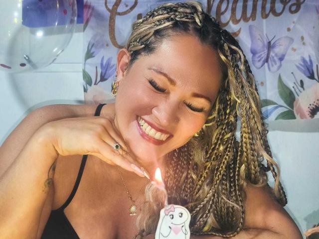 Vianca Castellanos, com cabelos loiros trançados, sorrindo, olhos fechados, segurando uma vela com um adesivo fofo, vestindo um vestido preto com alças, usando brincos de ouro, um anel e um colar, em um quarto bem iluminado..