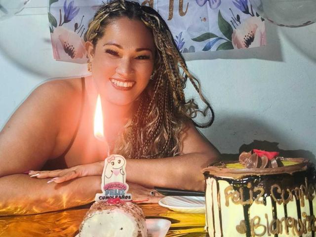 Vianca Castellanos, met krullend blond haar, lachend, met kaarslicht, zonder top, die een taart vasthoudt met een decoratie en een andere taart met "Happy Birthday" in chocolade. Borsten zichtbaar, vrolijke uitdrukking..