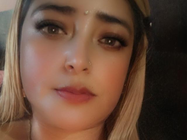 AlessadraJoness with blonde hair, dark eyebrows, brown eyes, subtle makeup, pink lipstick, nose piercing, and a small dot on her forehead, looking slightly confused.