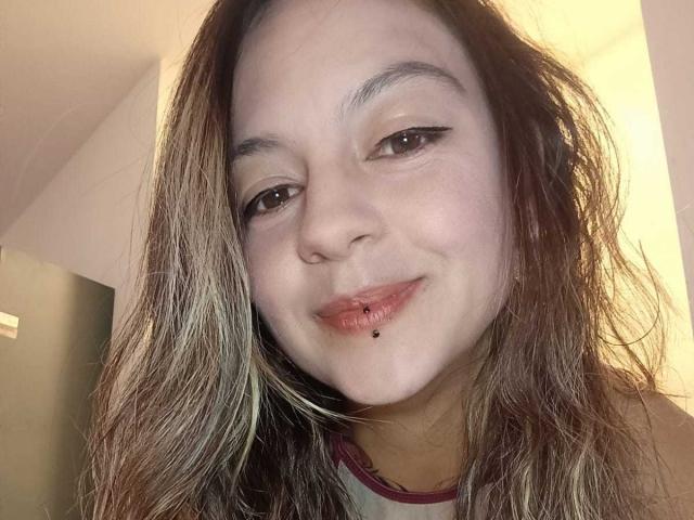 ArianaBlonde with a slight smirk, brown eyes, and a lip piercing, has messy blonde-brown hair, wearing a red-trimmed shirt. Her nose is small, and she's looking at the camera.
