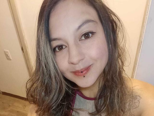 ArianaBlonde has wavy, dirty-blonde hair, brown eyes, and a slight smile with a lip piercing. She's wearing a grey top with pink trim.