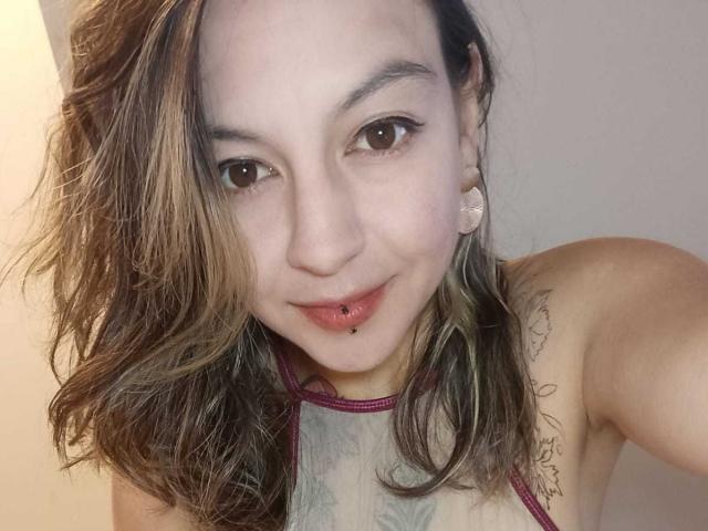 ArianaBlonde with wavy brown hair, light skin, brown eyes, and a lip piercing, wearing a maroon halter top, showing tattoos on her chest and shoulder.