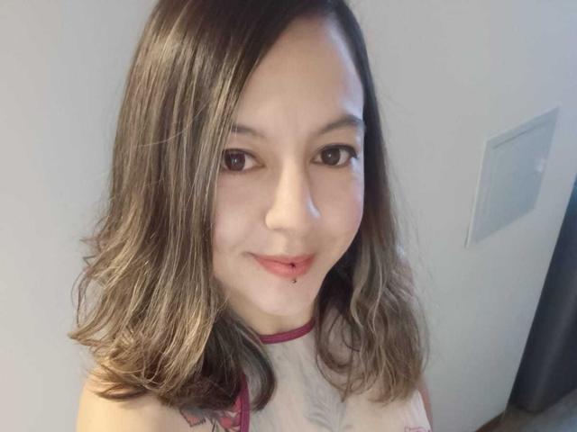 ArianaBlonde has light skin, brown eyes, and shoulder-length brown hair. She's wearing a white top with red trim, smiling, and has a small cut on her lower lip.