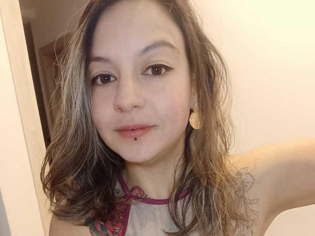 ArianaBlonde has wavy brown hair, light skin, and a small lip piercing. She's wearing a sleeveless top with a pink halter, showing her small tits.