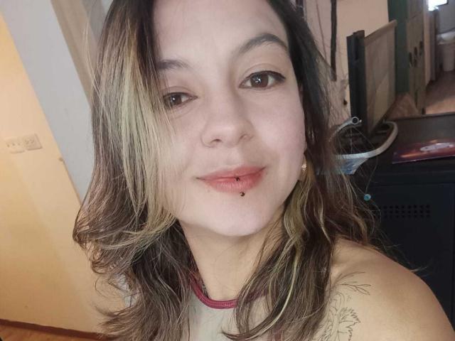 ArianaBlonde with medium brown hair, light pink lipstick, and a lip piercing, smiling at the camera. She has a tattoo on her right shoulder and is wearing a red tank top.