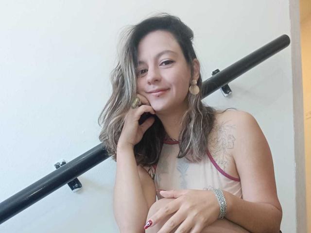 ArianaBlonde with wavy brown hair, small tits, tattoo on right shoulder, smiling, wearing sheer top with red trim, holding rail, earbuds in, silver bracelet, red nails.