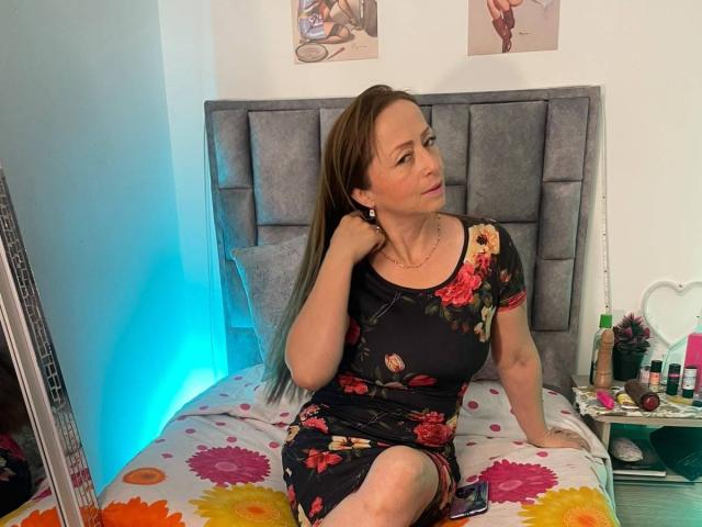 GianineLarose, with long brown hair, wearing a black floral dress, sits on a bed with colorful floral sheets, looking to the side, hand on her ear.