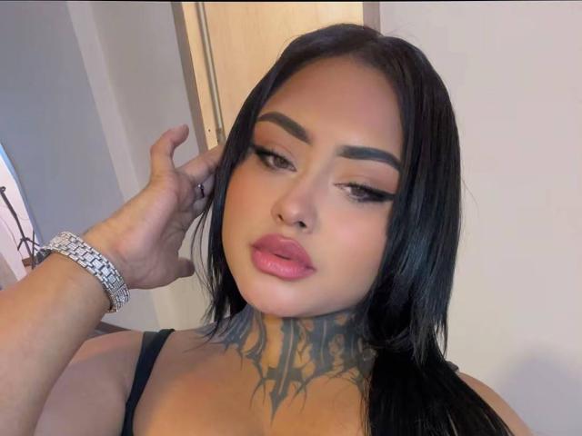 LairaHearvy with black hair, thick eyebrows, pink lips, and a tattoo on her neck, wearing a black strap, touching her hair, wearing a silver bracelet.