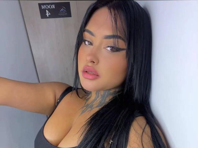 LairaHearvy has black hair, thick eyebrows, winged eyeliner, pink lips, tattoo on her neck, and large boobs in a black tank top, standing against a wall.