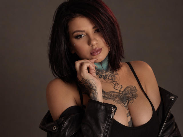 SophieWilds, com jaqueta de couro preta nos ombros, top preto que mostra seios grandes, peito e mão tatuados, cabelo curto castanho escuro com mechas vermelhas e colar azul..