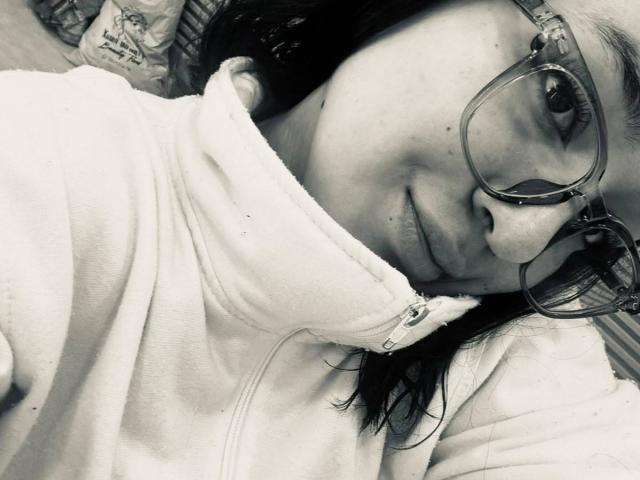NiniWillow with glasses, lying down, smiling slightly, wearing a zipped-up hoodie, black hair, fair skin, in a black-and-white photo, showing her nose and part of her mouth.