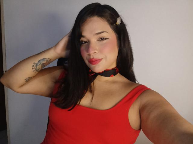 AntonelaPark with dark hair, red dress, black and red choker, tattoos on arm, hand in hair, smiling, pink blush, red lipstick, selfie, light skin, cute, sexy.