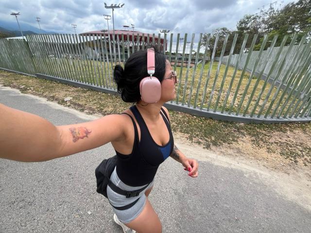 AlanaLuxe, wearing pink headphones, black tank top, gray shorts, and glasses, takes a selfie. She has a tattoo on her right arm and is outside with a metal fence.