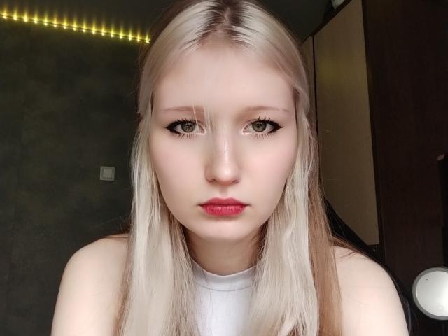 Deandrea Winkelbauer, con la pelle chiara, i capelli biondi lunghi, gli occhi verdi, il rossetto rosso e la maglietta bianca, guarda direttamente la fotocamera con un'espressione seria..