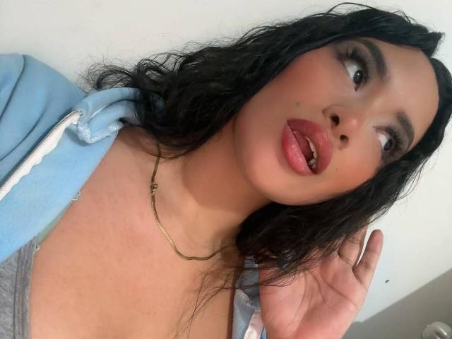 Adalina Vera, avec ses cheveux noirs mouillés, ses lèvres roses et pulpeuses, et un collier en or, porte un sweat à capuche bleu et un haut gris, et regarde vers le haut avec une bouche légèrement ouverte. Ses fesses ne sont pas visibles..