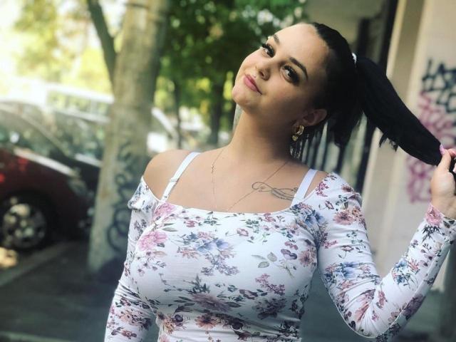 MilaFawn, avec de grandes seins, un haut à fleurs avec une épaule dénudée, et une queue de cheval haute, les cheveux noirs attachés, souriante, avec des tatouages sur le côté droit, des boucles d'oreilles dorées, debout à l'extérieur, une journée ensoleillée, un décor urbain..