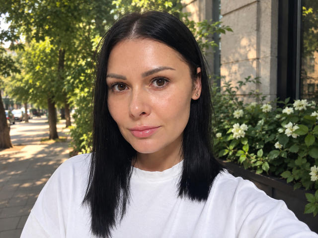 Ariana Diaz mit glattem, schwarzem Haar, braunen Augen und rosafarbenen Lippen, trägt ein weißes Hemd und steht draußen vor einem Grünflächenbereich und einem Gebäude. Sie schaut in die Kamera..