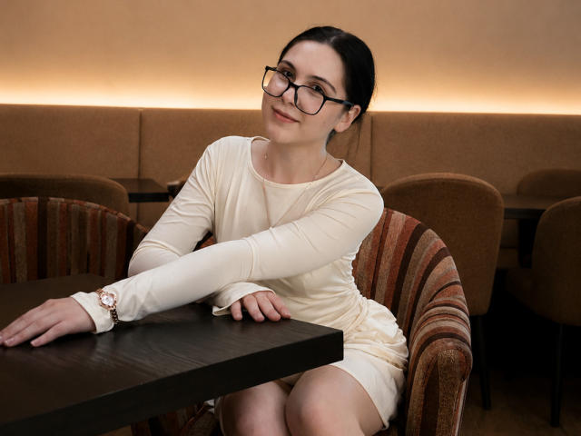 DeeannMilloy with black glasses, wearing a white long-sleeve dress, sitting at a table, leaning on it with one arm, smiling, brown chair with red stripes, wrist bracelet.