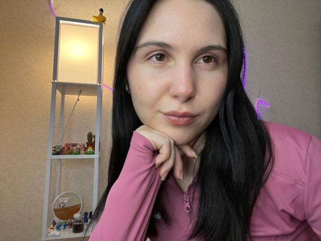 DeeannMilloy with long black hair, pink shirt, and pouty lips, resting her chin on her hand, brown eyes, slight smile, purple LED lights in the background.