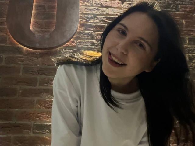 DeeannMilloy has black hair, a white shirt, and is smiling with her tongue slightly out, looking to the side in a dimly lit, brick-walled setting.