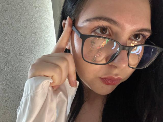 DeeannMilloy with glasses, holding shirt, black hair, brown eyes, reflection on glasses, slightly parted pink lips, white shirt, left hand on cheek, textured wall behind.