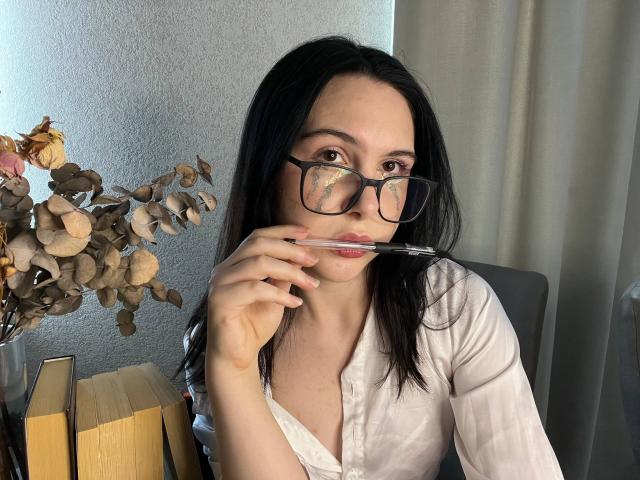 DeeannMilloy with black glasses, white shirt, and pink lipstick, holding a pen to her lips, has dark hair, and is sitting near a vase with dried flowers.