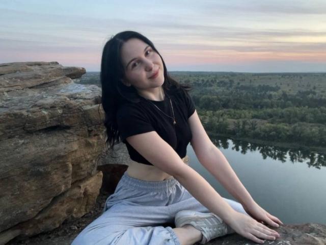 DeeannMilloy with dark hair, black crop top, gray pants, sitting on rocky ledge, hands on rock, subtle smile, sunset sky, reflection in water, forest in background.