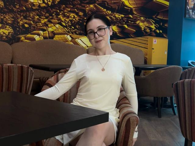 DeeannMilloy in a white dress, sitting in a brown armchair, wearing glasses, with a necklace, and a small ass, in a cafe with wooden tables and striped chairs.