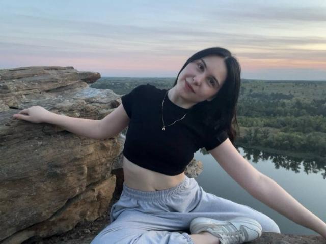 DeeannMilloy with black hair, small tits, wearing a black crop top and gray sweatpants, sitting on rocky ledge, smiling at sunset, arm resting on rock, necklace visible.