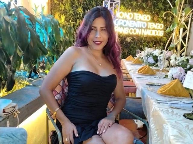 LunaTomson with purple-dyed hair, wearing a strapless black dress, sitting at a table with yellow napkins, and white floral decorations, showing her tits and smiling.