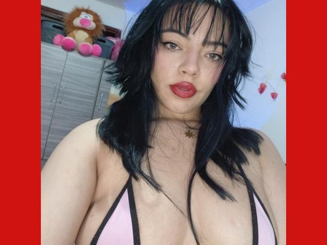 AbbieAllenn with black hair, pink bikini top, showing her big tits, red lipstick, and a star necklace, standing in a room with a stuffed lion. Red border.