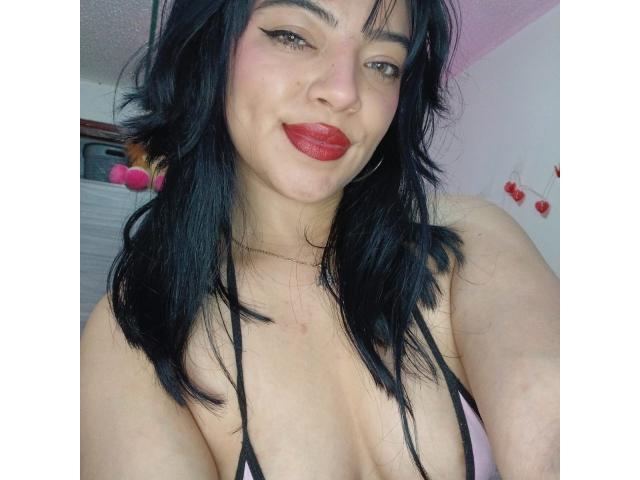 AbbieAllenn with black hair, red lipstick, and black strappy top showing her tits, smiling slightly, with a small scar on her chest. She's wearing hoop earrings.
