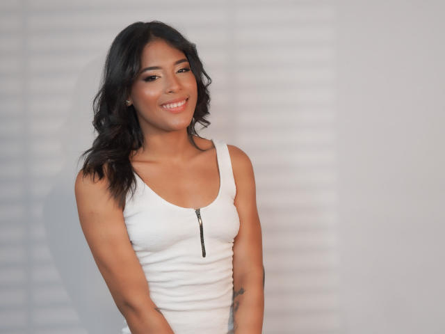 AgataDolly with dark wavy hair, wearing a white tank top with a zipper, smiling, showing her small tits and a tattoo on her left arm, in a photo.