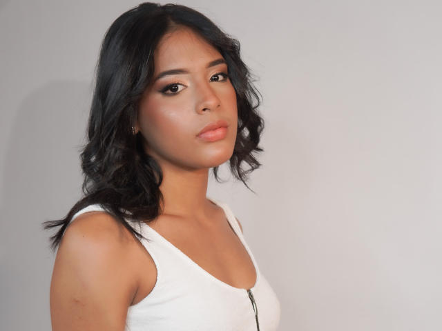 AgataDolly has medium brown skin, black wavy hair, and is wearing a white tank top with a zipper. Her boobs are small, and she's looking serious.