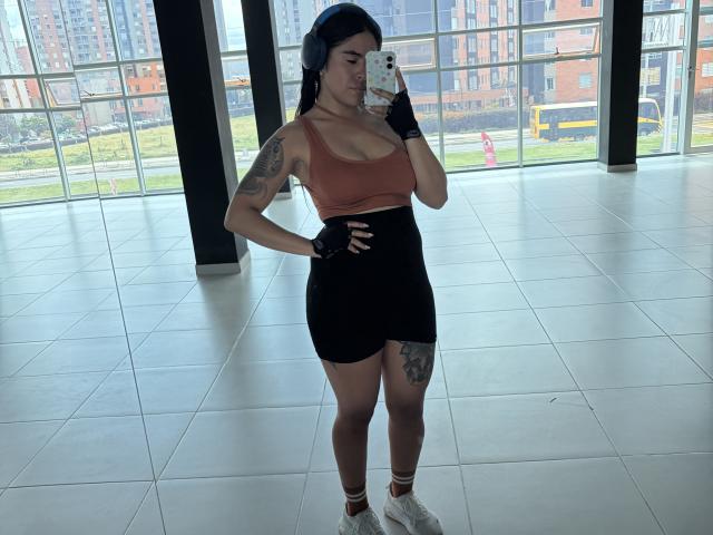 StellaBeckker has big boobs, a tattooed arm, and thigh, wearing a brown sports bra, black shorts, finger gloves, and white socks with sneakers, taking a selfie in a glass building.