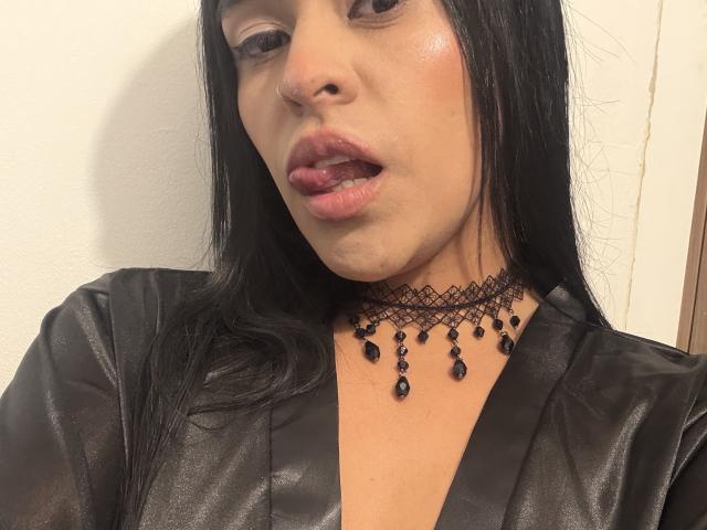 StellaBeckker has long black hair, shiny skin, and a black lace choker with teardrop beads. She's wearing a black satin robe, with her tongue slightly out.