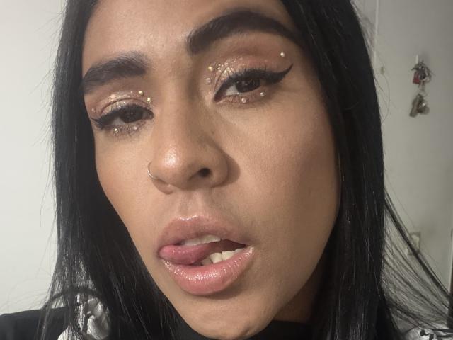 Le visage de StellaBeckker, avec ses lèvres roses et brillantes légèrement entrouvertes, révélant un eye-liner et des cils dorés, un piercing au nez, et de longs cheveux noirs, regarde directement l'appareil photo..