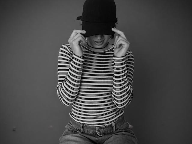 NaraStorn, die ein gestreiftes Hemd und Jeans trägt, zieht eine dunkel gefärbte Baseballkappe über ihren Kopf und lächelt leicht. Schwarz-weiß-Foto..