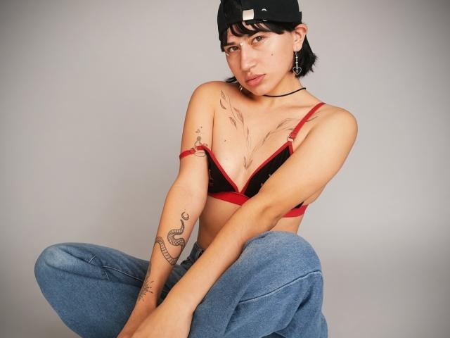 NaraStorn with short black hair, black cap, red bra, blue jeans, sitting cross-legged, tattoos on arms, choker necklace, looking at camera, subtle expression, light skin, small tits.
