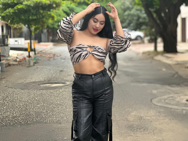MiaBallard with long black hair, zebra-print off-shoulder top, and black leather pants, standing on a wet street, hands in hair, looking down, red lipstick.