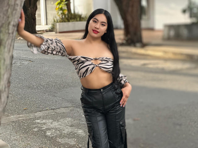 Mia Ballard, avec ses longs cheveux noirs, son rouge à lèvres rouge, son haut à rayures zébrées qui dévoile ses seins, et son pantalon en cuir noir, appuyée contre un arbre, une main sur la hanche, un sourire confiant..