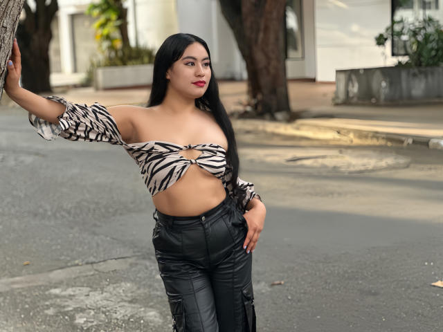 MiaBallard with long black hair, zebra print off-shoulder top exposing her small tits, black leather pants, standing outside, right arm on tree, looking to the side, red lips.