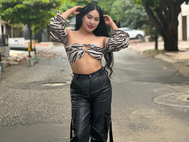 Mia Ballard mit langen, schwarzen Haaren, einem zebrastreifenartigen Top, das ihre Brüste entblößt, schwarzen Lederhosen, stehend auf einer nassen Straße, Hände im Haar, lächelnd, rote Lippenstift..