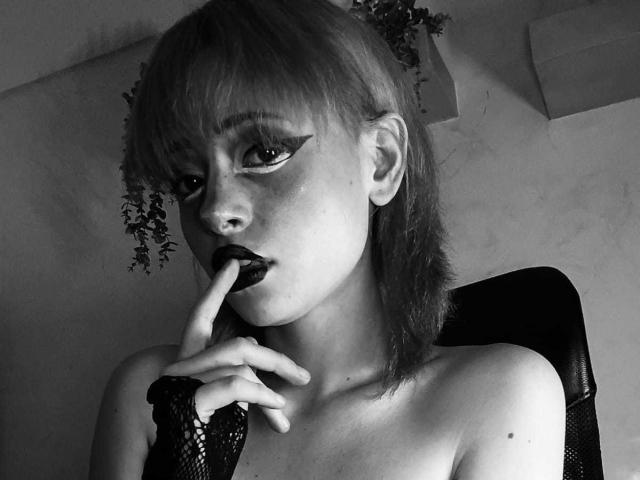 ViolettClay with pale skin, dark lipstick, and heavy eyeliner, biting her finger, wearing a black mesh glove, and no top, with short blonde hair and a pensive expression.