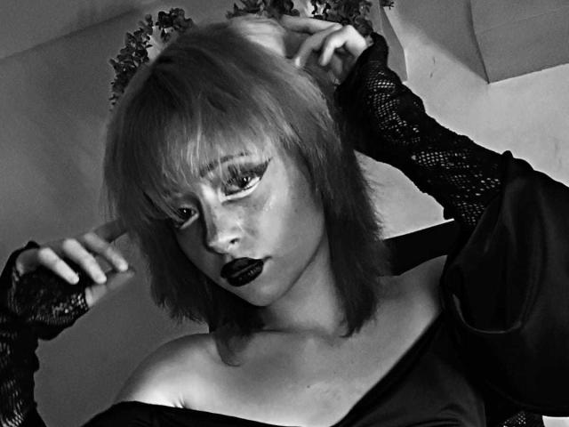 ViolettClay has goth makeup, dark lipstick, and lace sleeves, holding her short, blonde hair with a floral headpiece, wearing an off-shoulder top, in a black-and-white photo.