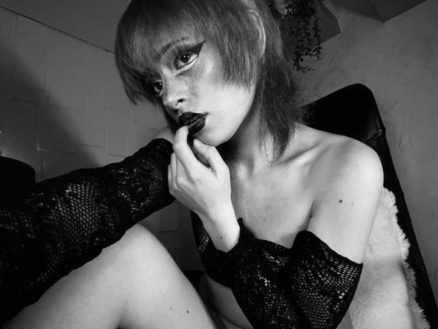 ViolettClay with dark makeup, biting a black candy, wearing a fishnet off-shoulder top, exposing her small tits and fair skin, with short, messy hair, in a black-and-white shot.