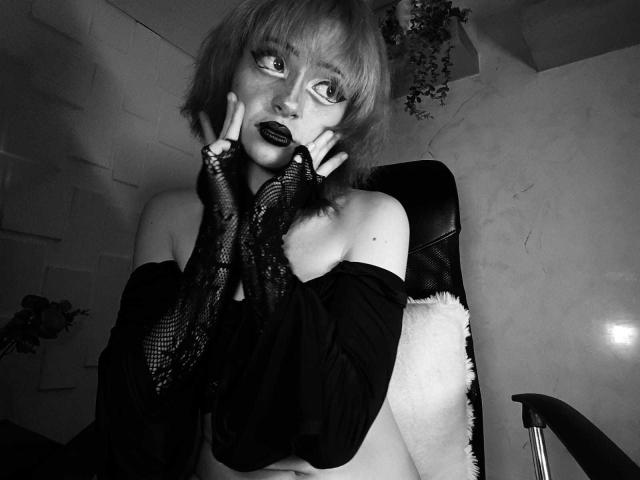 Black-and-white photo of ViolettClay with short, light-colored hair, wearing fishnet gloves, off-shoulder top, making double-v sign near face, showing dark lipstick and freckles, sitting on chair.