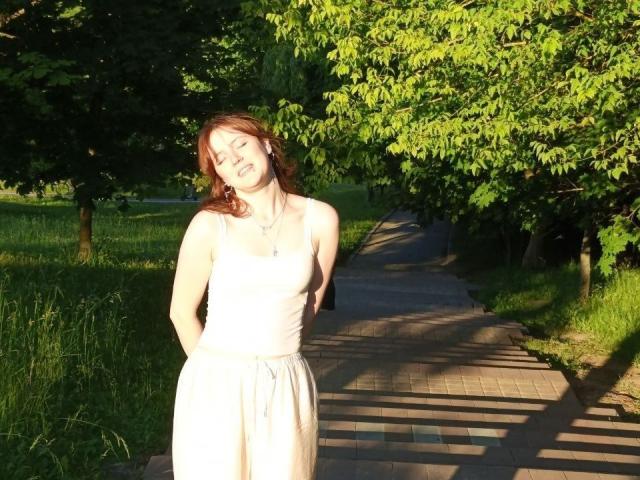 PatSanabria está de pie en un camino iluminado por el sol, con los ojos cerrados, sonriendo y con las manos detrás de la espalda. Lleva un vestido blanco, un collar y tiene el pelo rojo. Árboles y hierba verdes la rodean..