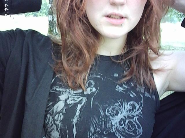 PatSanabria with red hair, pale skin, and a black graphic tank top showing a detailed wolf pattern, has her arm raised and a slightly open mouth.
