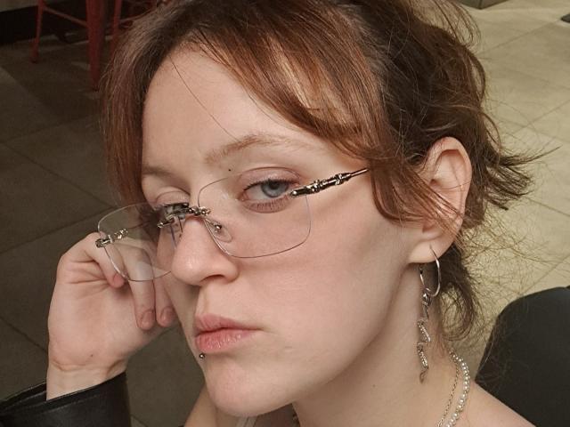 PatSanabria has pale skin, brown hair in a messy updo, wearing glasses with black zippers, a nose piercing, and a lip piercing, with a necklace and dangling earrings.